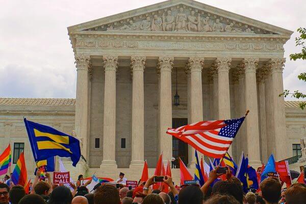 A crowd in front of the supreme court holding flags for gay pride and equality. There are signs reading "love for all" and "equality forward."