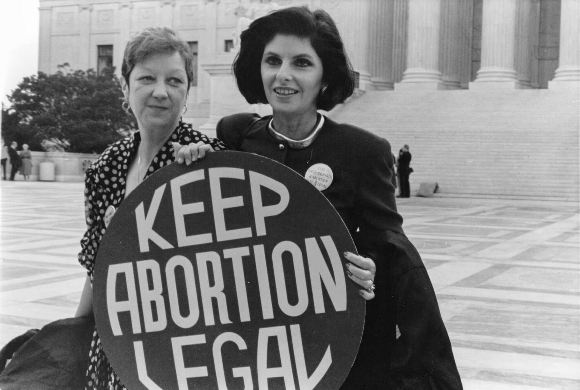 Two women standing in front of the supreme court building holding a sign that says "keep abortion legal."