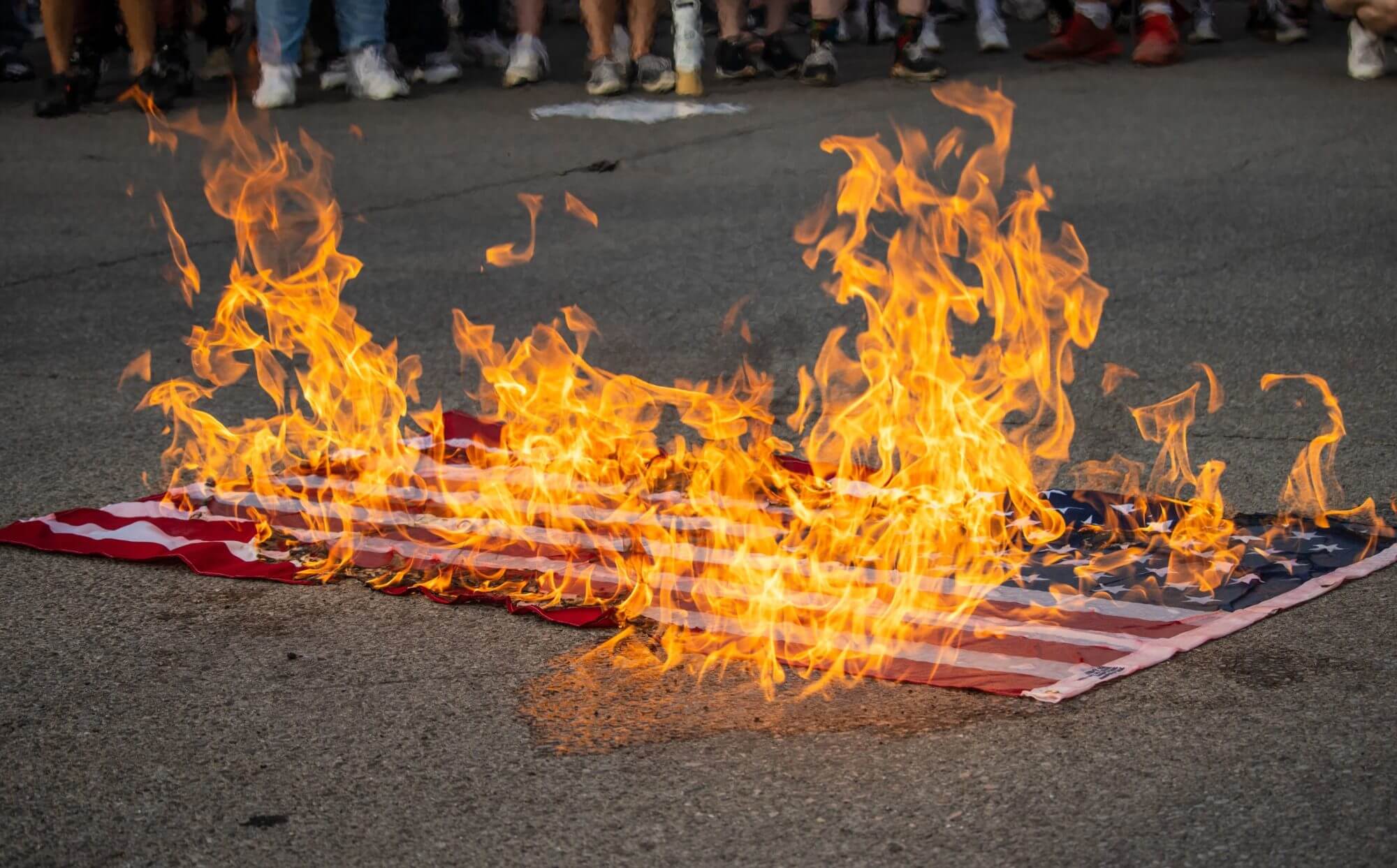 An American flag burning in the street in the middle of a crowd.